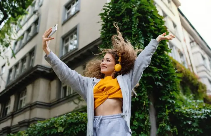 Smiling young woman with headphones raising her arms and holding a smartphone on a city street