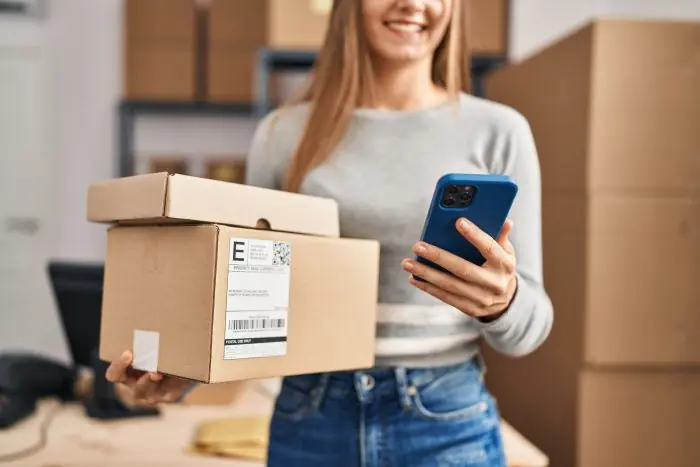 Smiling woman holding a cardboard package and a smartphone in a shipping office