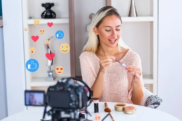 Woman applying makeup in front of a camera while social media like and emoji icons float beside her