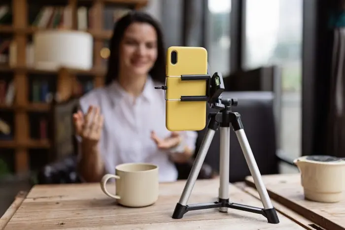 Woman gesturing at a smartphone mounted on a tripod to record a video, with a coffee cup on the table