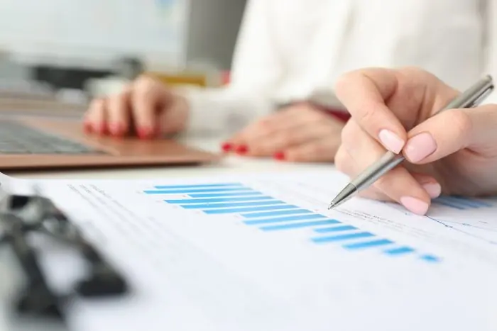 Hand holding a pen over a printed bar chart while another person types on a laptop