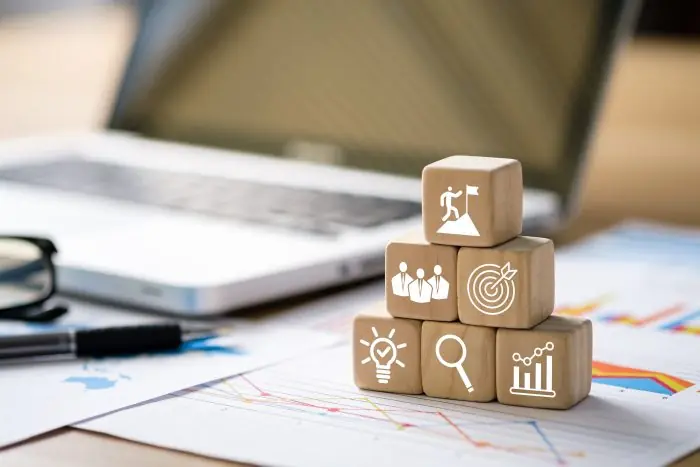 Wooden blocks with goal, team, target, idea, search and growth icons stacked on desk with charts and laptop
