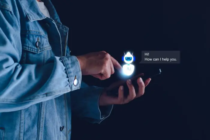Hand tapping a smartphone with a glowing AI chatbot icon saying 'Hi! How can I help you.'
