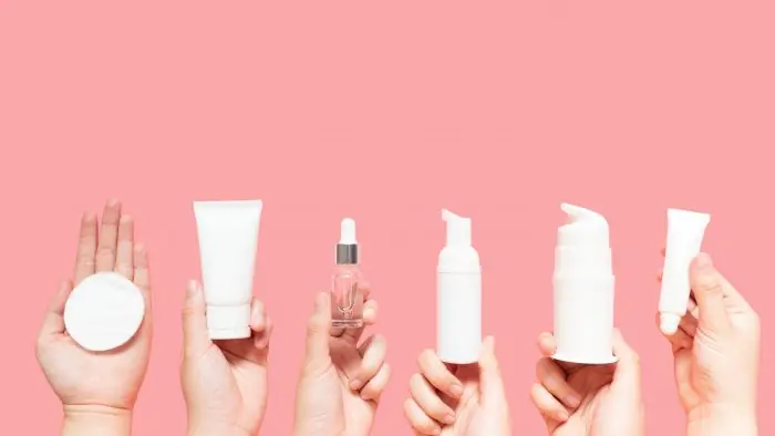 Six hands holding white skincare products and a cotton pad against a pink background