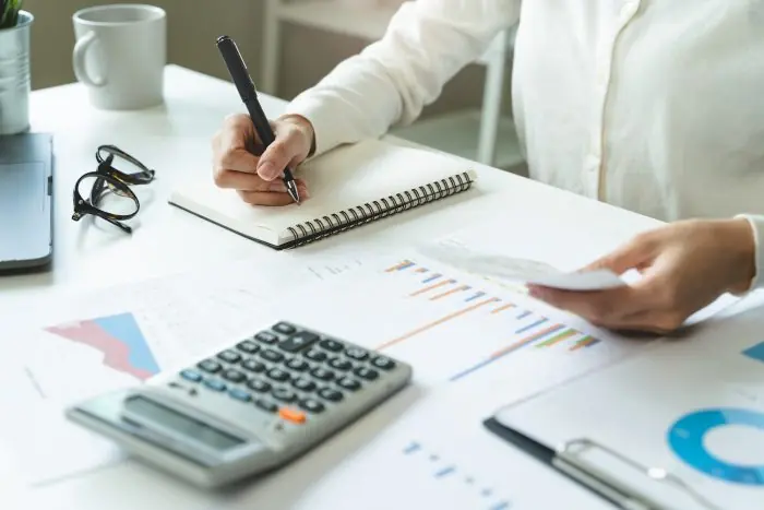 Person writing in a notebook at a desk with a calculator, glasses and printed charts