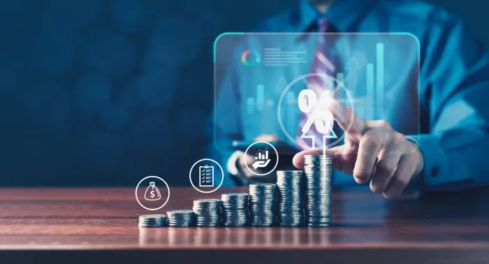 Hand touching a percent symbol above stacked coins and financial icons on a holographic chart, representing growth
