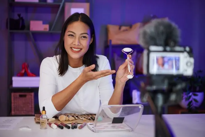 Smiling woman holding a facial roller and presenting makeup products in front of a camera