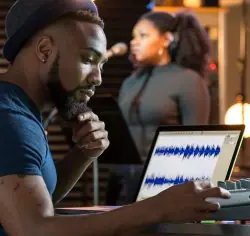 Man listening with a hand on his chin while viewing an audio waveform on a laptop, with a woman behind him